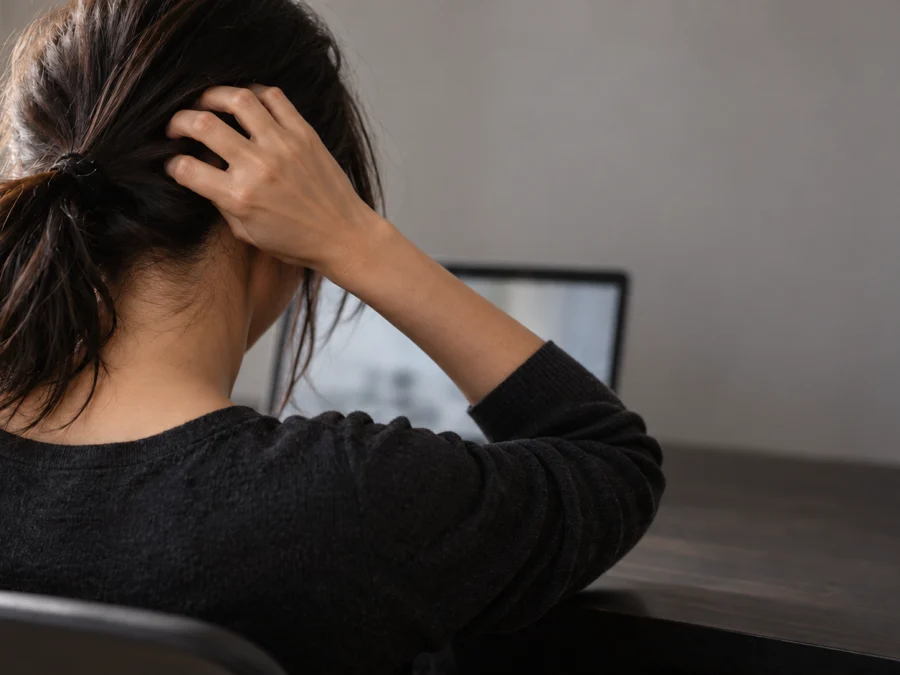 Itchy scalp and stress: woman seen from behind at a desk with a laptop, dark hair in a low ponytail, fingers scratching the scalp at the back of the head — SIMONE TRICHOLOGY