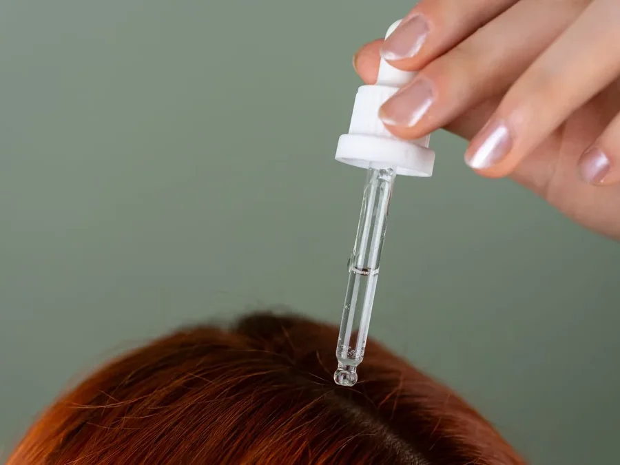 Hand applying a clear serum with a dropper to the roots of red hair as part of a scalp care routine