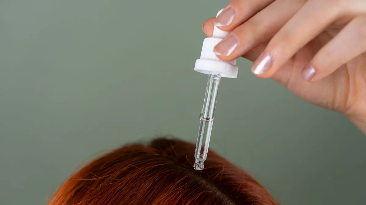 Hand applying a clear serum with a dropper to the roots of red hair as part of a scalp care routine